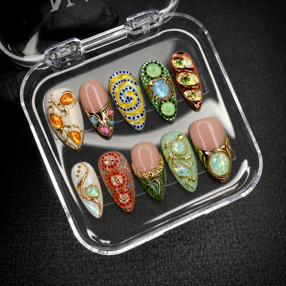 Open clear nail case on a dark textured surface holding ten coordinated nails with gold-tone ornament, colorful patterns, and iridescent-looking accents.