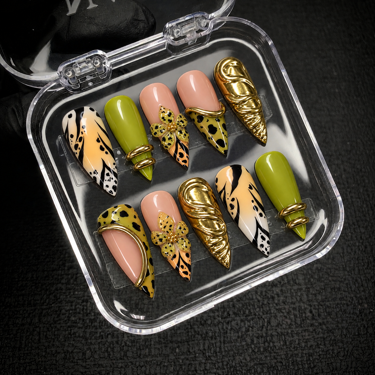 Open clear case on dark textured fabric displaying ten coordinated nails with chartreuse green, spotted flowers, black markings, and gold-tone accents.