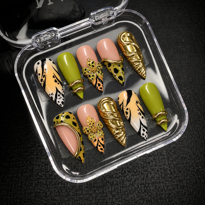 Open clear case on dark textured fabric displaying ten coordinated nails with chartreuse green, spotted flowers, black markings, and gold-tone accents.