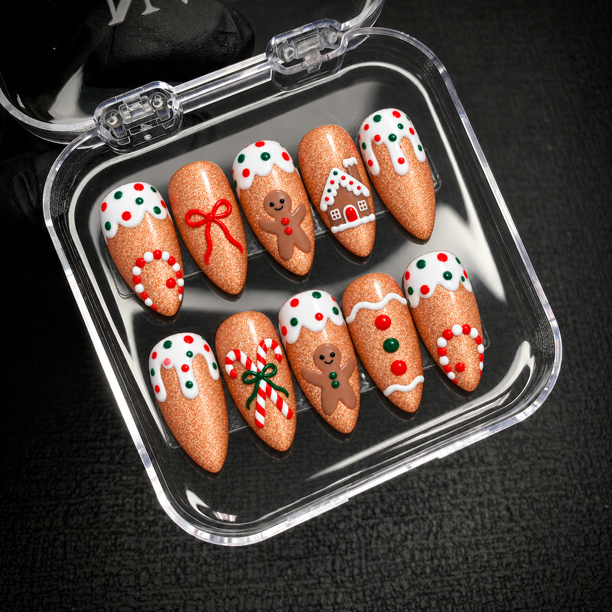 Open clear nail case on dark textured fabric displaying ten caramel-brown nails with gingerbread figures, white icing, candy canes, and festive red-and-green details.