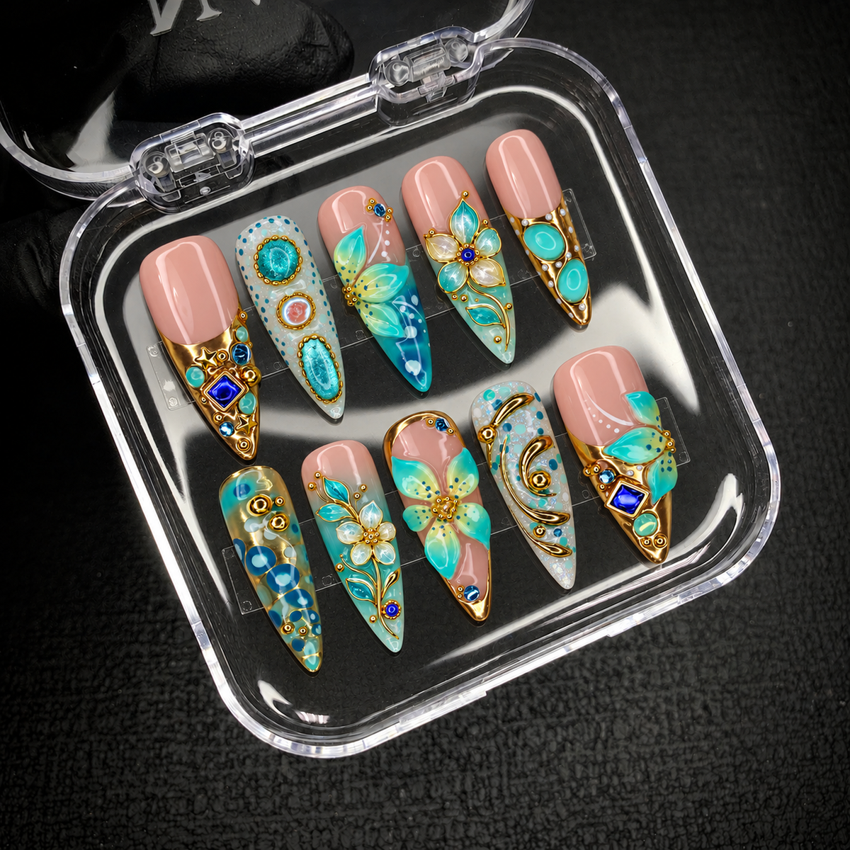 Open clear display case holding ten coordinated nails with turquoise florals, gold-tone details, and cobalt accents against a dark textured background.