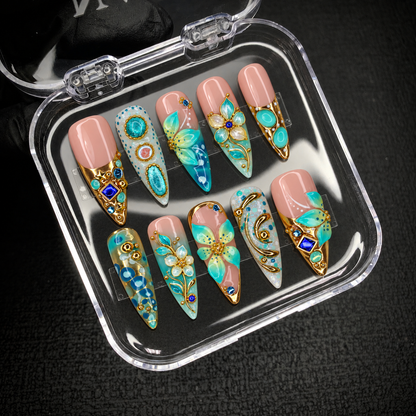 Open clear display case holding ten coordinated nails with turquoise florals, gold-tone details, and cobalt accents against a dark textured background.