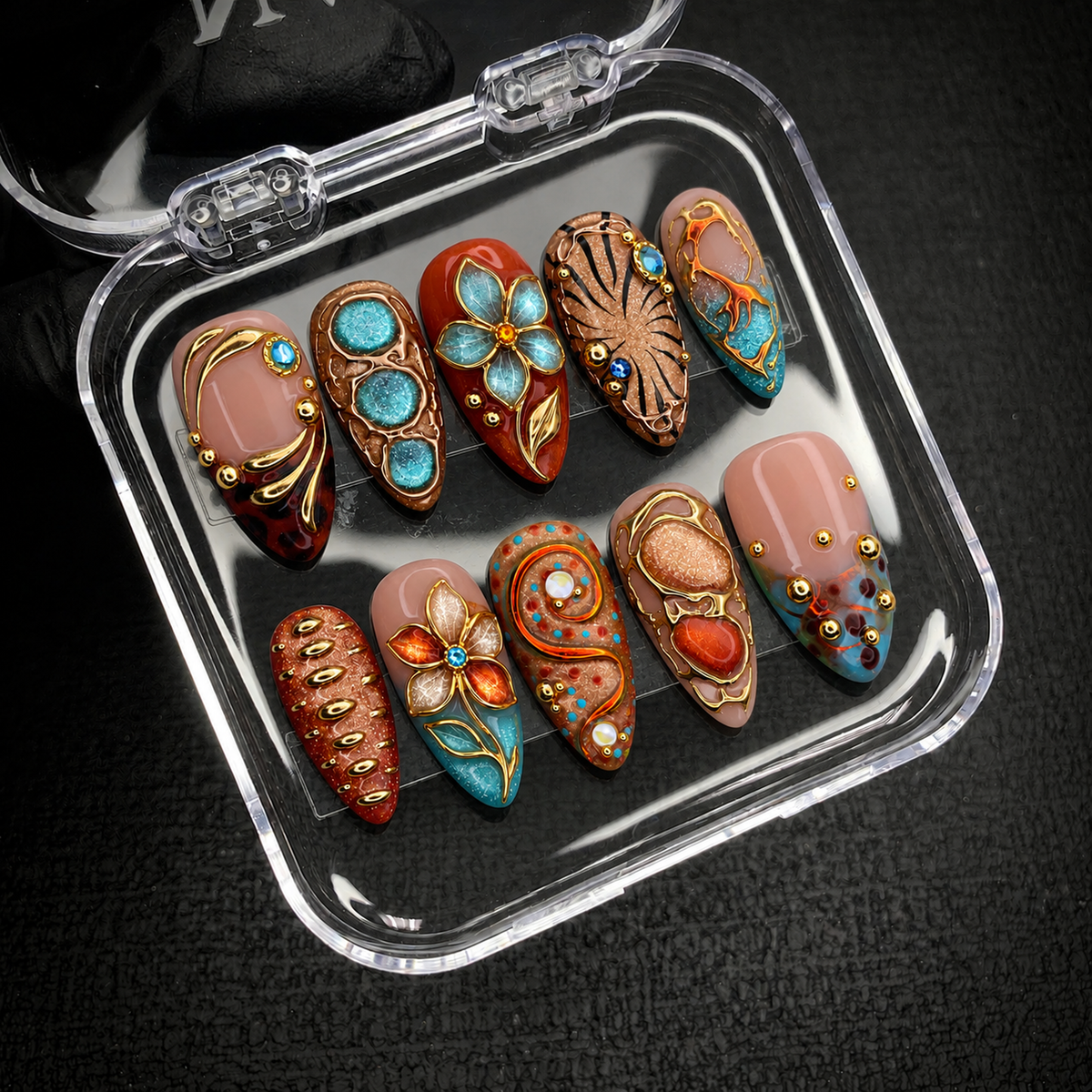 Open clear case holding ten coordinated turquoise, blush, and amber nails with ornate gold-colored accents on a dark textured background.