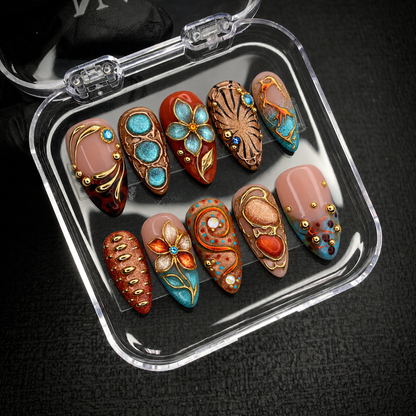 Open clear case holding ten coordinated turquoise, blush, and amber nails with ornate gold-colored accents on a dark textured background.