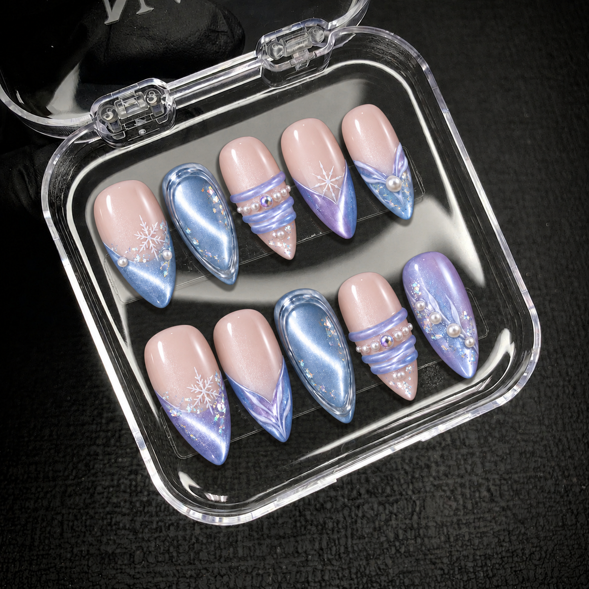 Open clear nail case on dark textured fabric holds ten coordinated blue, lilac, and blush-pink nails with snowflakes and raised embellishments.