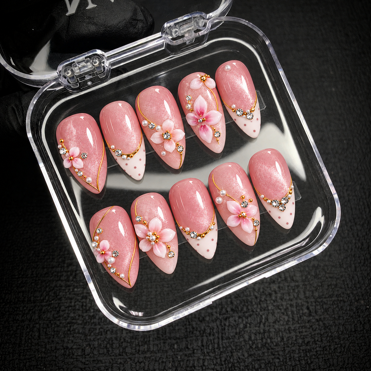 Open clear case on dark textured fabric holding ten blush pink nails with raised flowers, pearly accents, gold-colored lines, and dotted ivory tips.