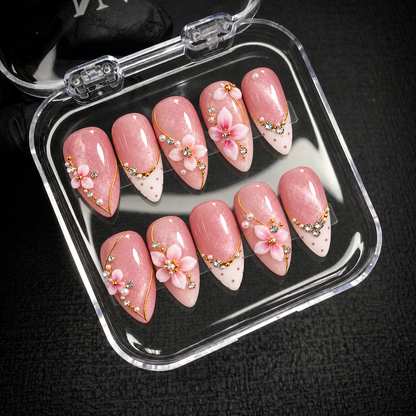 Open clear case on dark textured fabric holding ten blush pink nails with raised flowers, pearly accents, gold-colored lines, and dotted ivory tips.