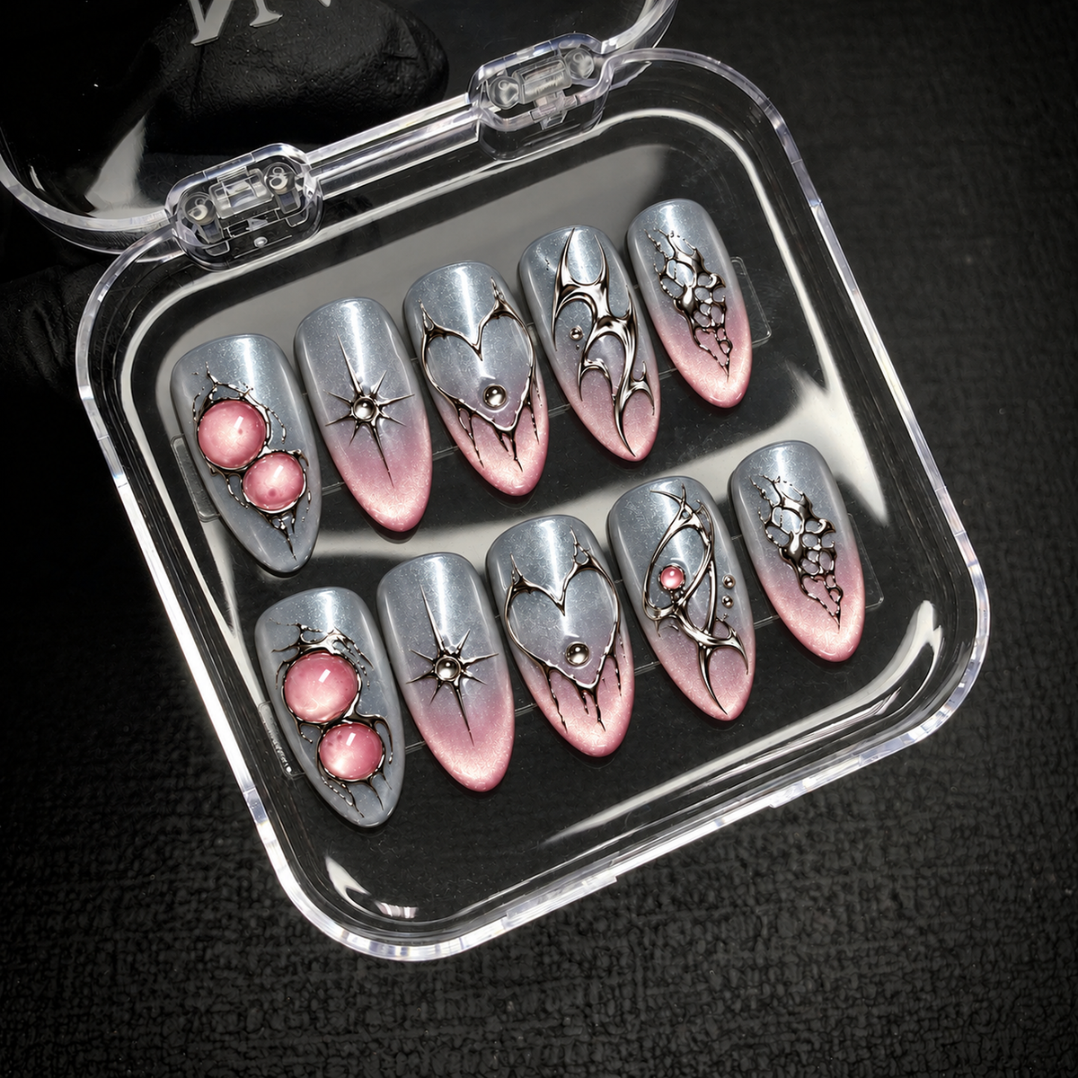 Open clear case holding ten silver-gray and blush pink nails with reflective ornamental details, positioned diagonally on dark textured fabric.