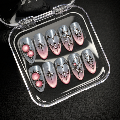 Open clear case holding ten silver-gray and blush pink nails with reflective ornamental details, positioned diagonally on dark textured fabric.