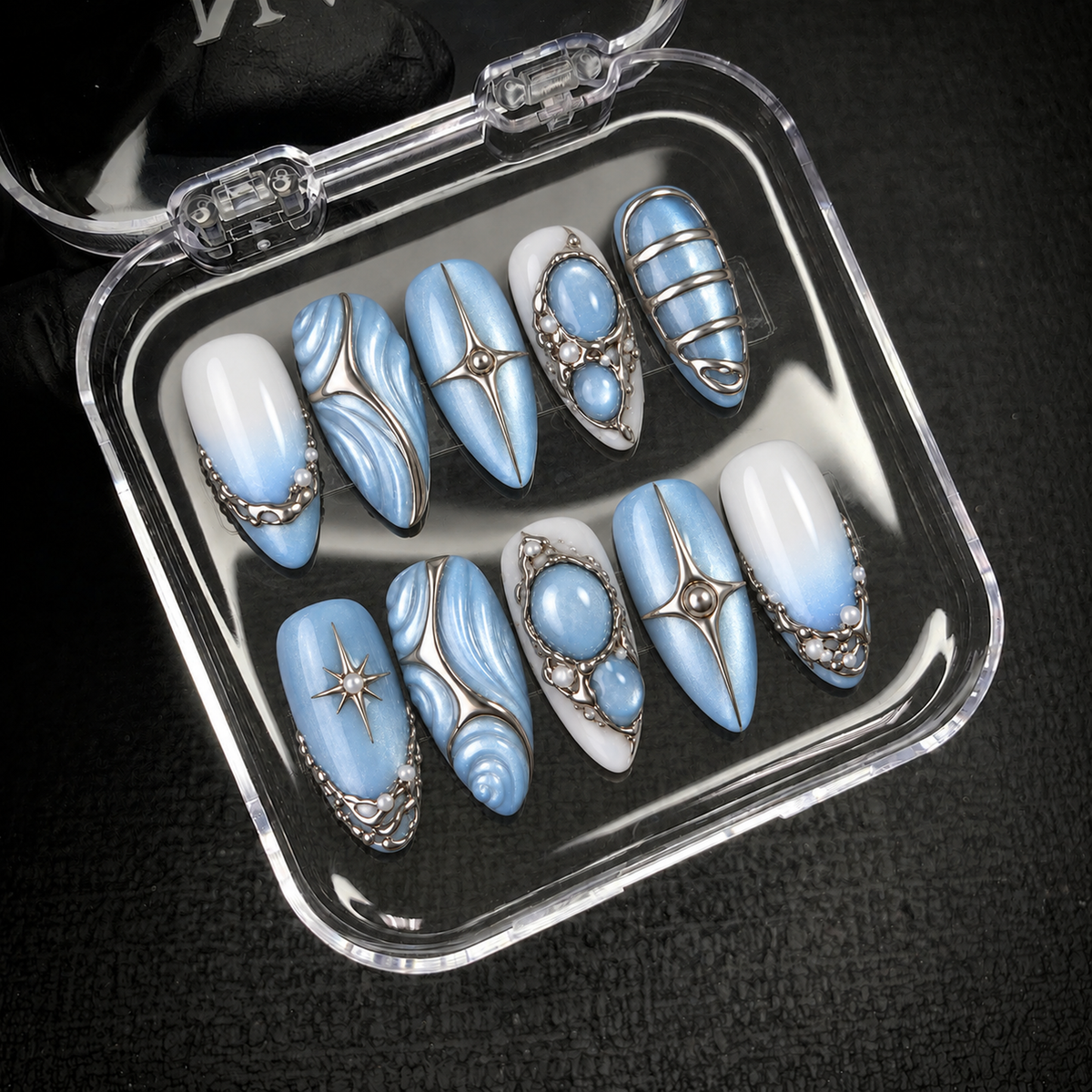 Ten blue and white Frosted Blue Tides nails arranged inside an open clear case, with silver-tone swirls, stars, bands, and oval accents visible.