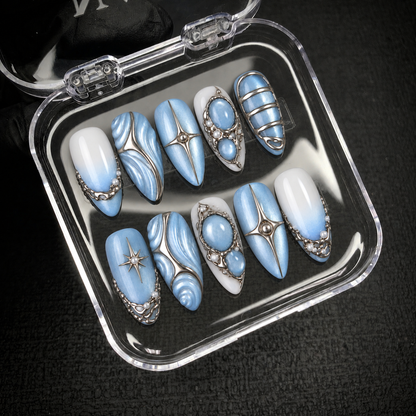 Ten blue and white Frosted Blue Tides nails arranged inside an open clear case, with silver-tone swirls, stars, bands, and oval accents visible.