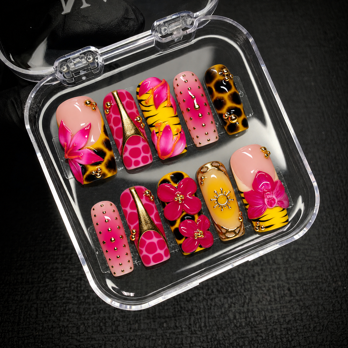 Open clear case on dark textured fabric displaying ten coordinated nails with pink blooms, animal patterns, gold-toned beads, and a sun accent.
