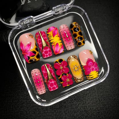 Open clear case on dark textured fabric displaying ten coordinated nails with pink blooms, animal patterns, gold-toned beads, and a sun accent.