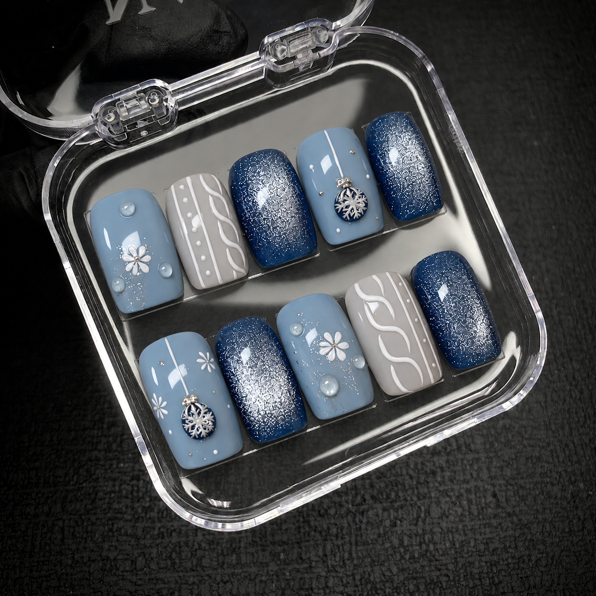 Open clear nail case on a dark textured surface displaying ten dusty blue, navy shimmer, and gray nails with floral, knit-inspired, and snowflake ornament details.