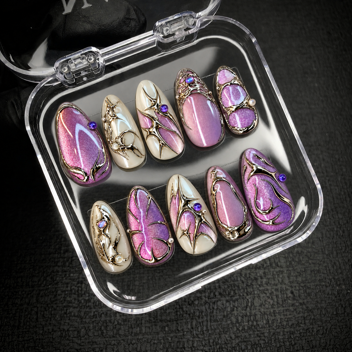 Open clear case holding ten pink-lilac and ivory nails with metallic filigree, set diagonally on a dark textured background.