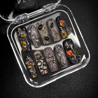 Open clear case holding ten coordinated smoky gray and charcoal nails with amber-orange accents and ornate details, displayed diagonally on a dark textured surface.