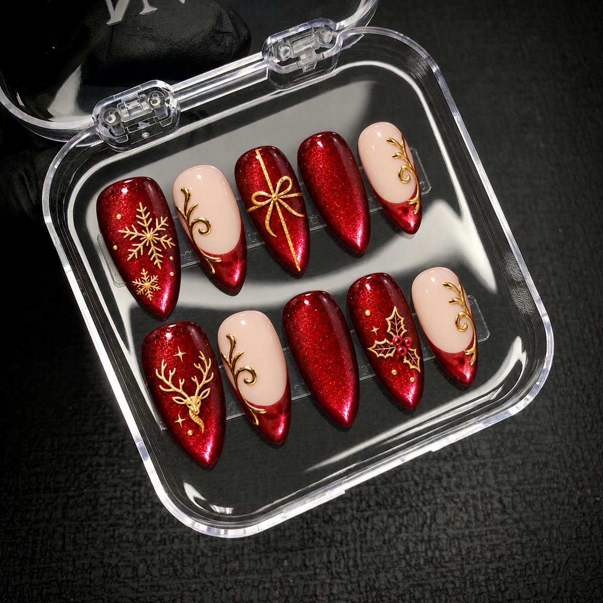 Open clear case holding ten coordinated ruby-red and blush Christmas nails with gold-tone decorations on a dark textured background.