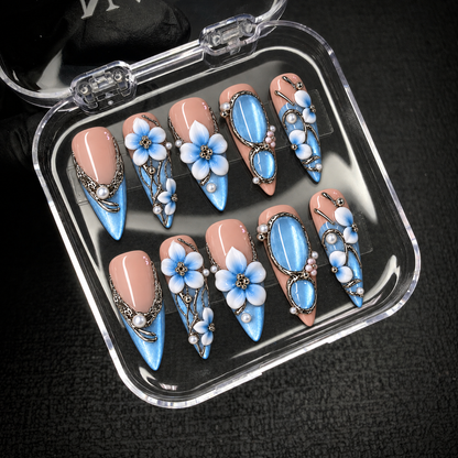 Open clear presentation box on a dark surface holding ten coordinated blush and blue floral press-on nails.