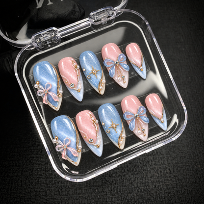 Open clear nail case on a dark textured surface holding ten coordinated pink and icy blue nails with ribbon bows, gold-tone details, and pearl-like accents.