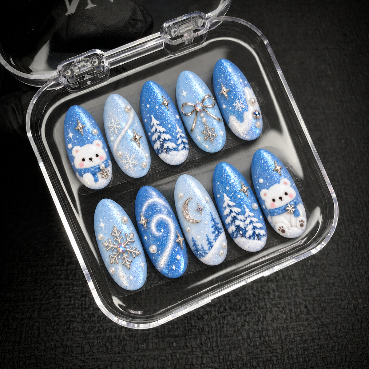 Open clear case on dark textured fabric holding ten blue winter nails with white bears, snowy evergreen trees, pearl-like accents, and silver-tone decorations.