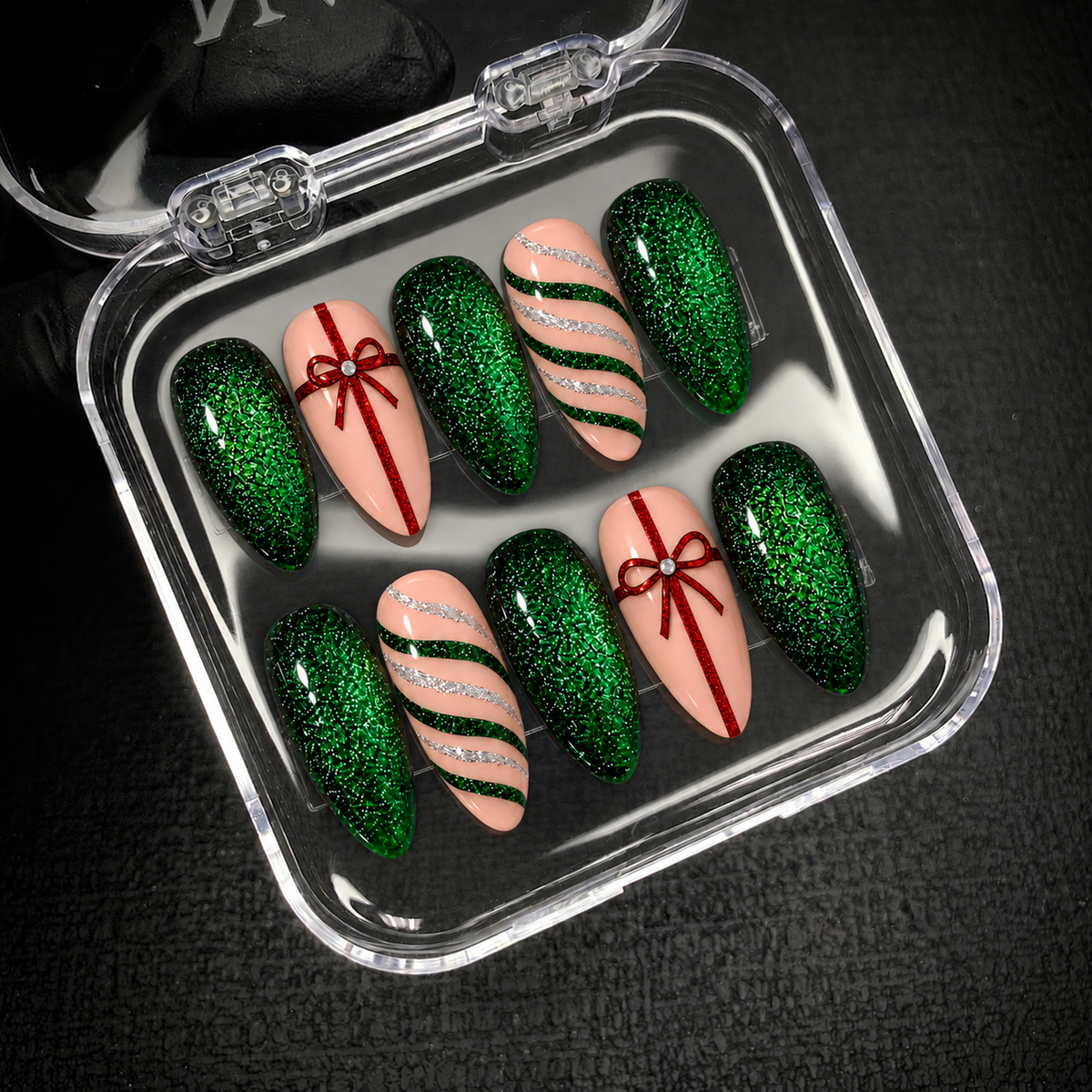 Open clear nail case on a dark textured surface holding ten emerald glitter and blush pink nails with red bows and green and silver stripes.