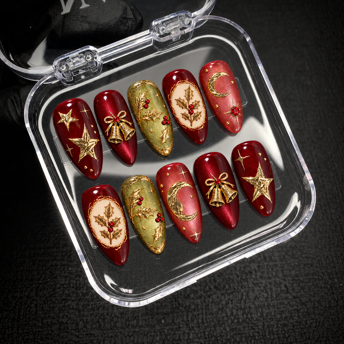 Open clear case on dark textured fabric displaying ten holiday press-on nails with gold-tone stars, bells, holly leaves, and crescent moons.