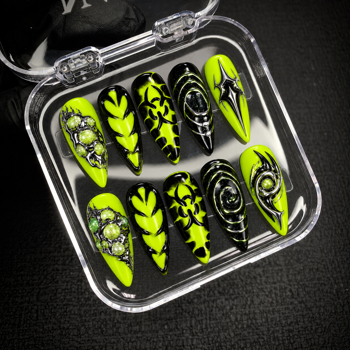 Open clear nail case on dark textured fabric displaying ten coordinated neon green and black nails with orbital graphics and silver-toned accents.