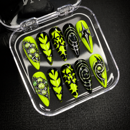 Open clear nail case on dark textured fabric displaying ten coordinated neon green and black nails with orbital graphics and silver-toned accents.
