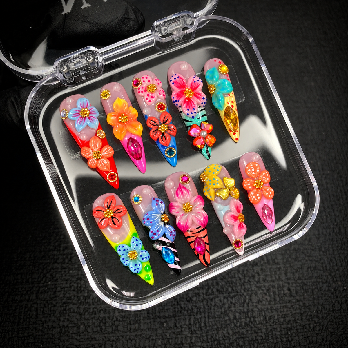 Open clear case holding ten colorful floral nails in two rows on a dark textured surface, showing raised flowers, striped sections, and faceted accents.