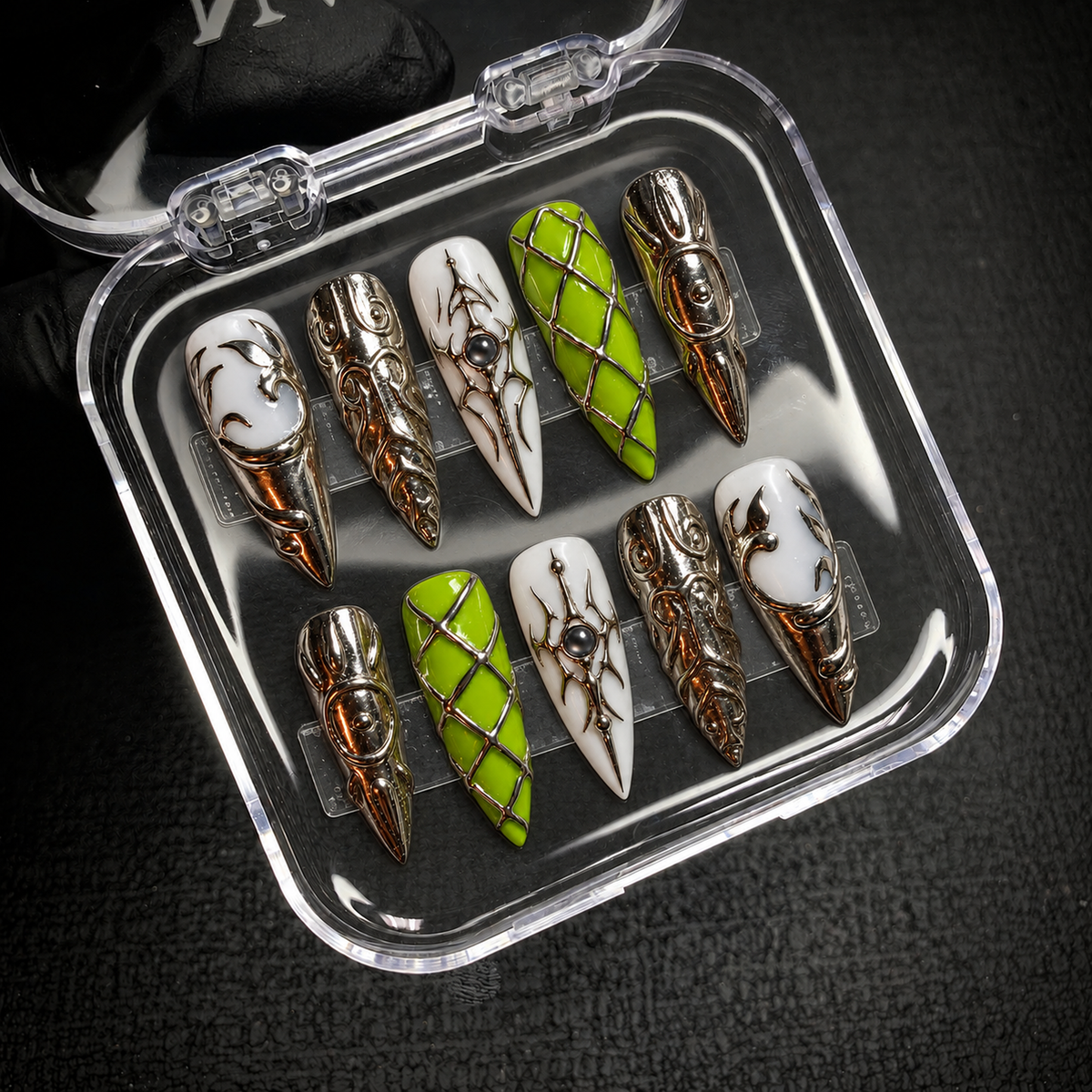 Open clear nail case on a dark textured surface holding ten coordinated lime green, milky white, and silver-tone nails with lattice and scroll motifs.