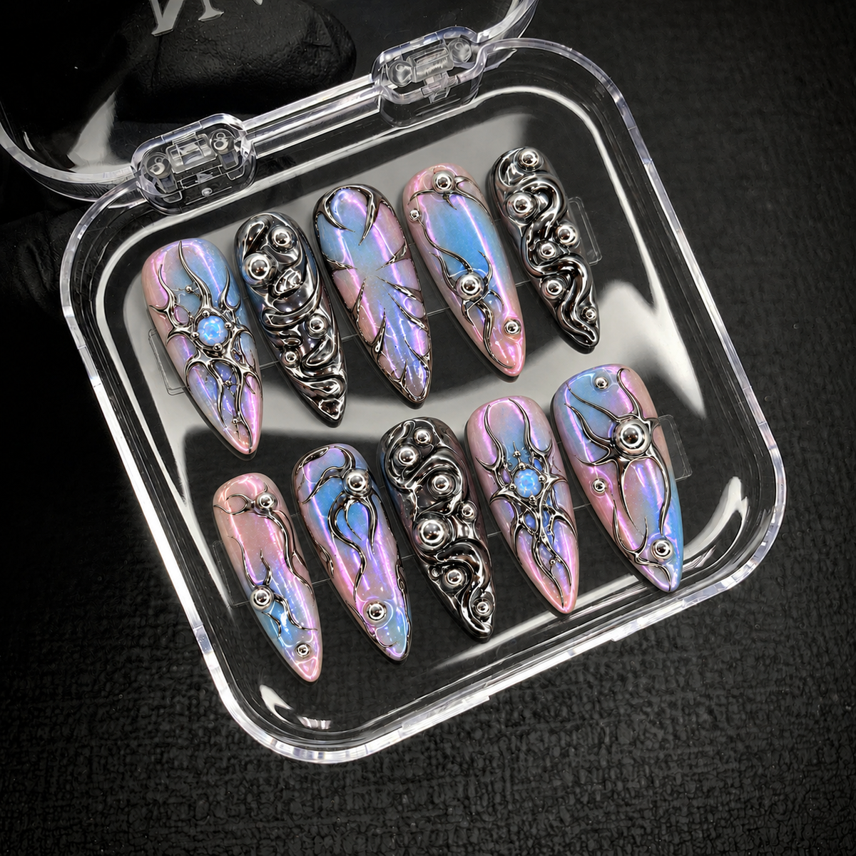 Open clear nail case on a dark textured surface holding ten coordinated nails with pastel iridescence, silver swirls, and opal-like blue details.