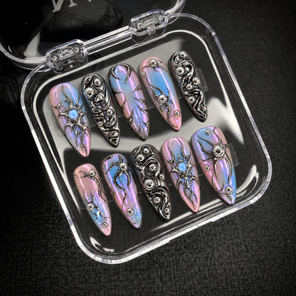 Open clear nail case on a dark textured surface holding ten coordinated nails with pastel iridescence, silver swirls, and opal-like blue details.