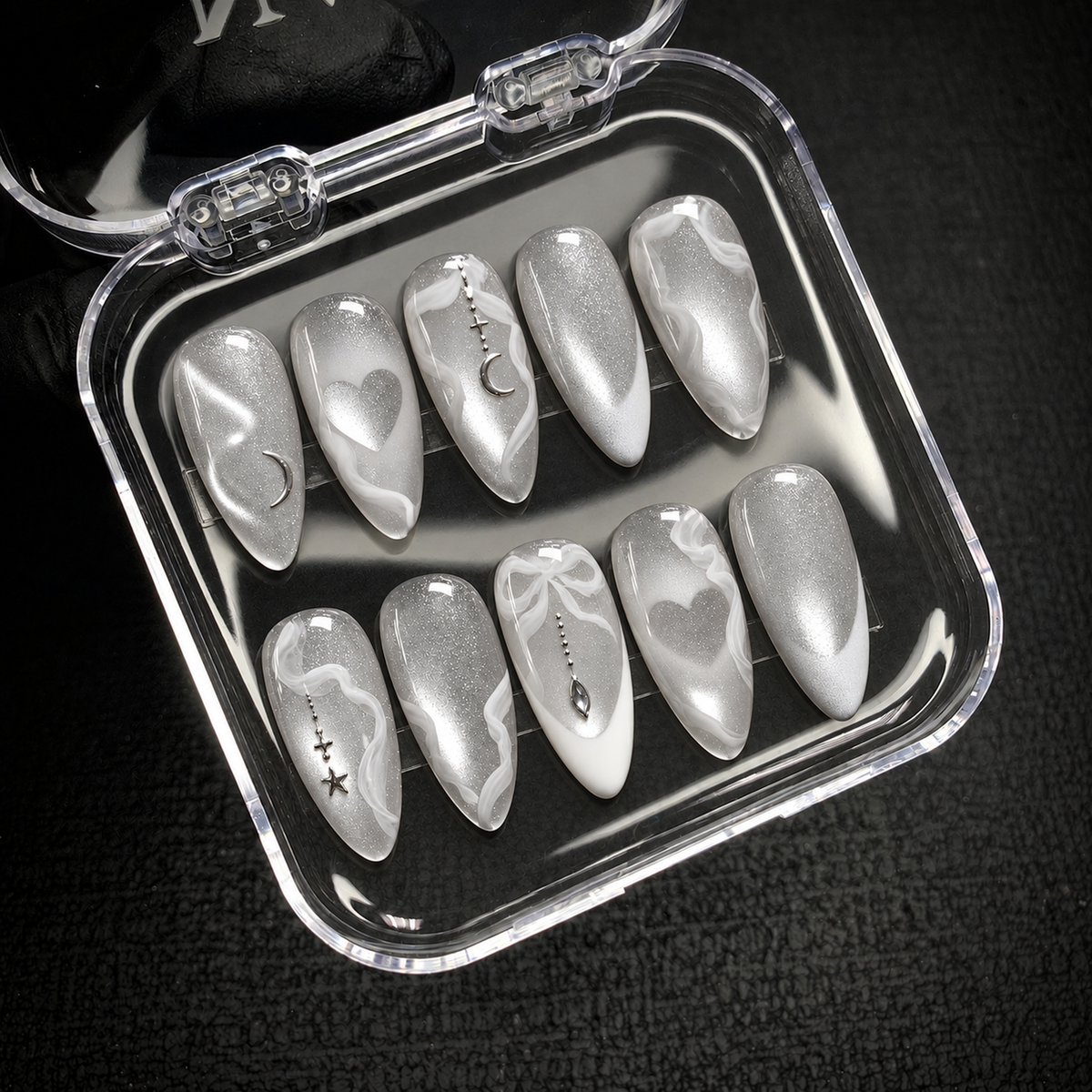 Open clear nail case on a dark textured surface displaying ten silver shimmer nails with white swirls and celestial and heart accents.