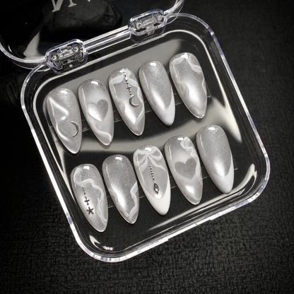 Open clear nail case on a dark textured surface displaying ten silver shimmer nails with white swirls and celestial and heart accents.
