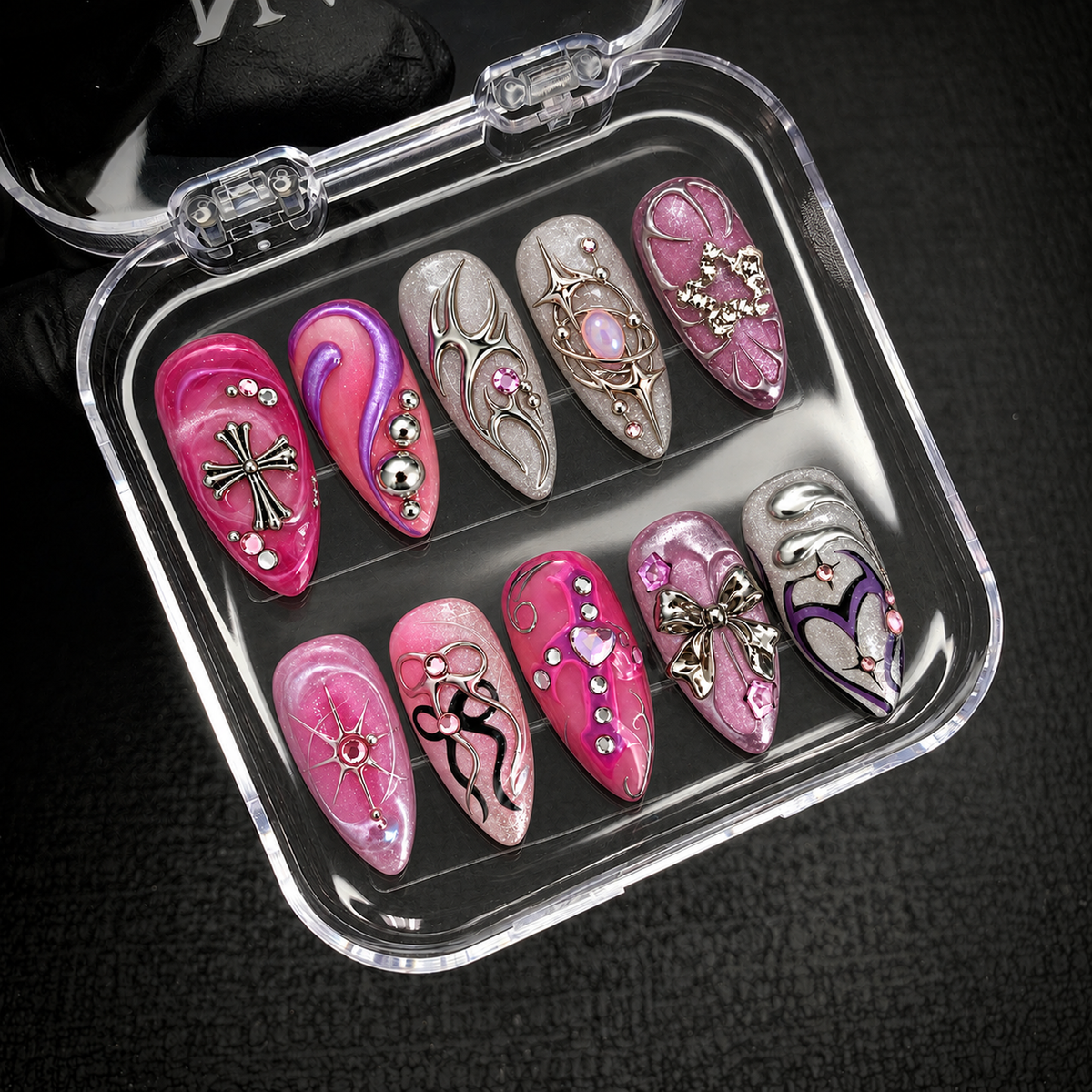 Open clear case holding ten pink, mauve, lilac, and silver decorated nails in two rows against a dark textured background.