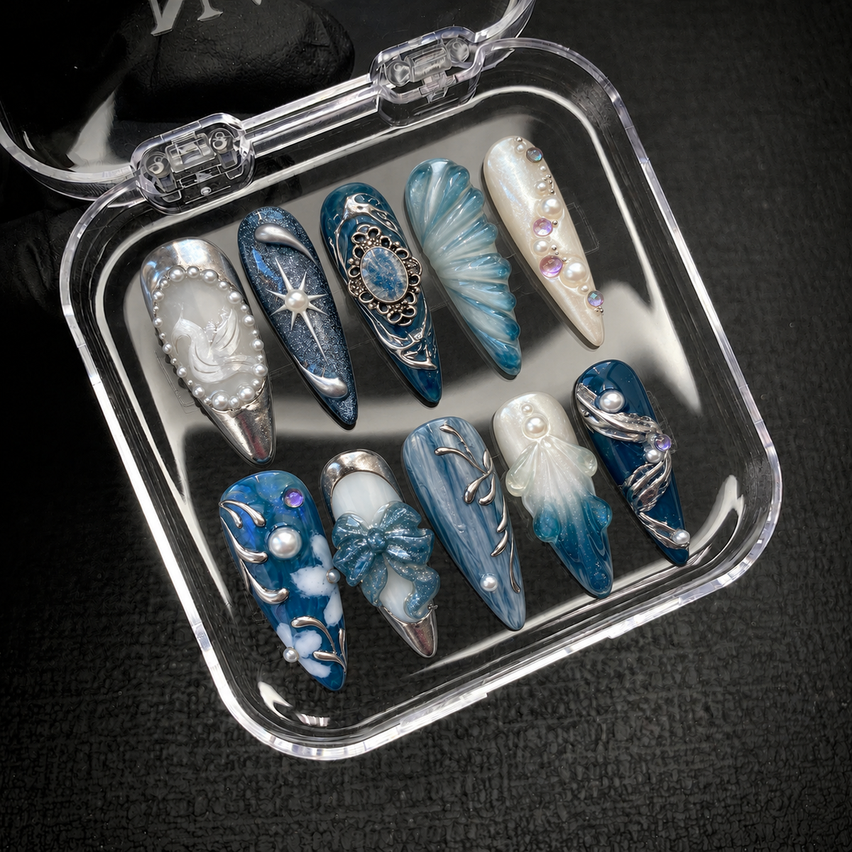 Open clear nail case on dark textured fabric displaying ten coordinated blue and pearl white nails with raised motifs and silver accents.