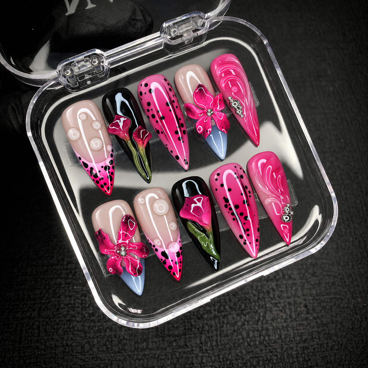 Open clear case holding ten coordinated pink, black, and nude floral nails, photographed at an angle on a dark textured surface.
