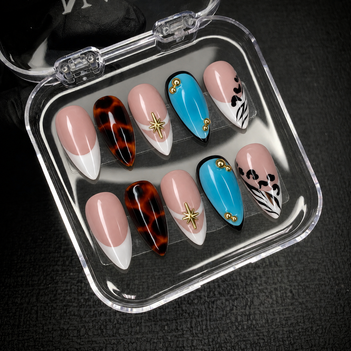 Open clear nail case on dark fabric displaying ten coordinated nails with turquoise accents, amber tortoiseshell, nude French tips, and gold-tone decorations.