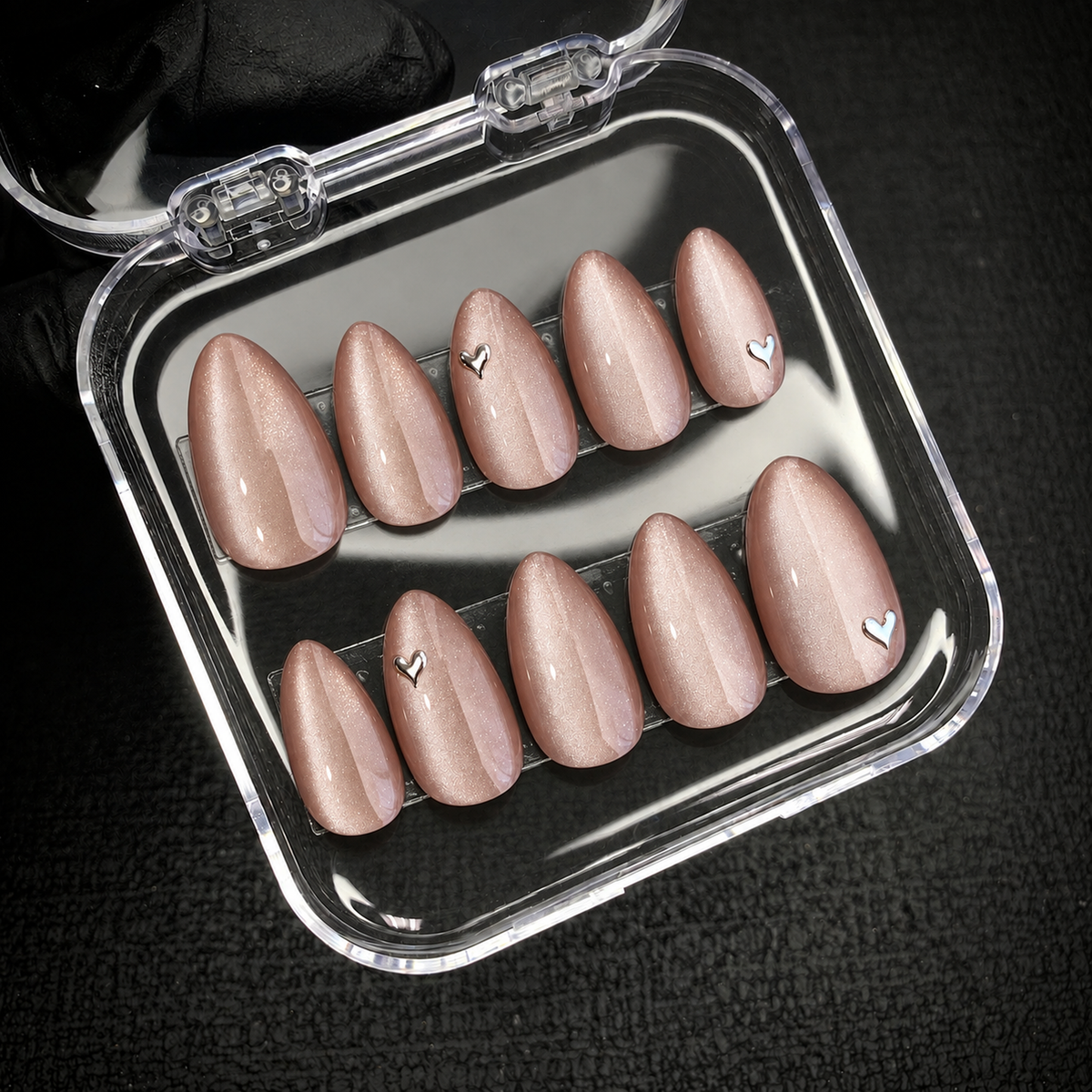 Open clear case holding ten blush-nude shimmer nails in two rows, including four silver-toned heart accents, against a dark textured background.
