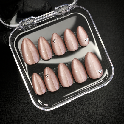 Open clear case holding ten blush-nude shimmer nails in two rows, including four silver-toned heart accents, against a dark textured background.