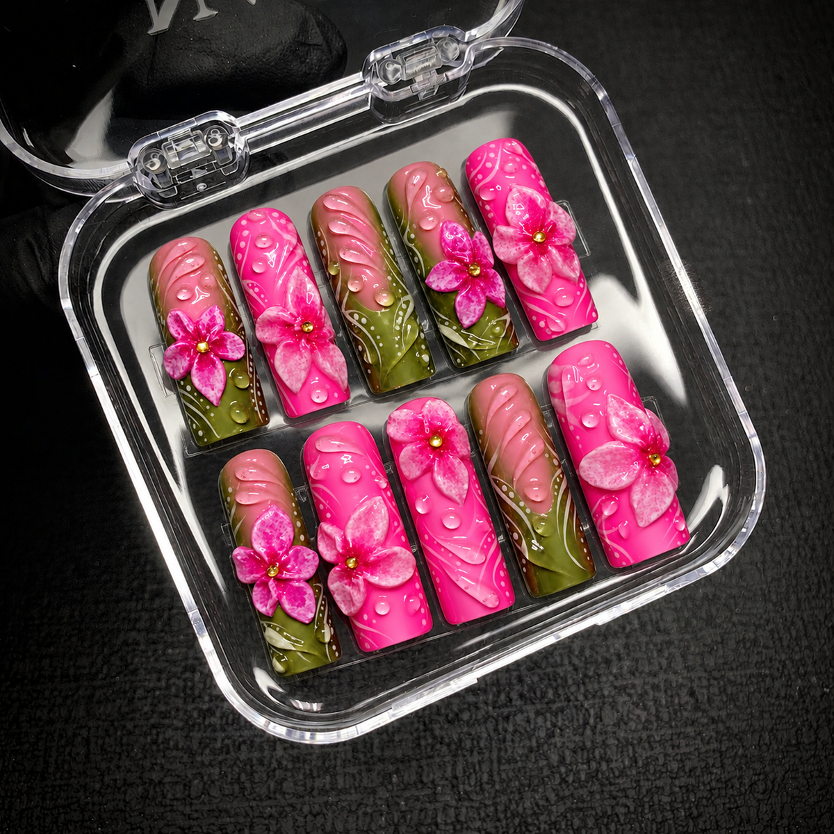 Open clear case holding ten pink and olive green floral nails in two rows on a dark textured background.