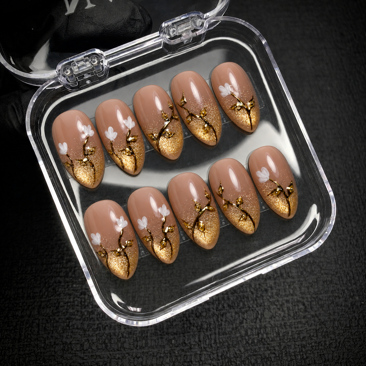 Open clear nail case on a dark textured surface holding ten nude nails with gold shimmer, branching leaf details, and white floral accents.