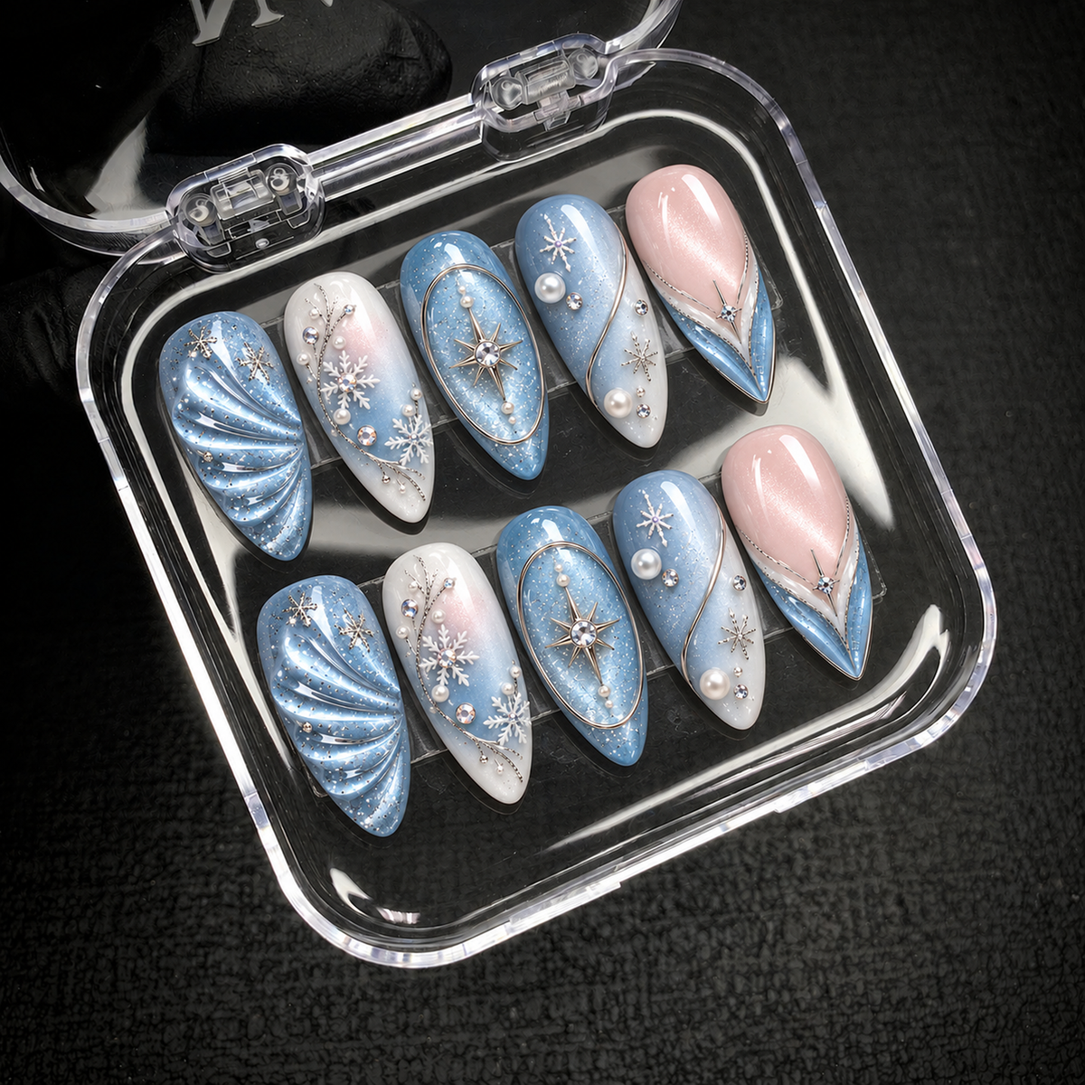 Open clear case holding ten coordinated winter nails with icy blue shimmer, blush pink accents, snowflakes, and starbursts on a dark textured surface.