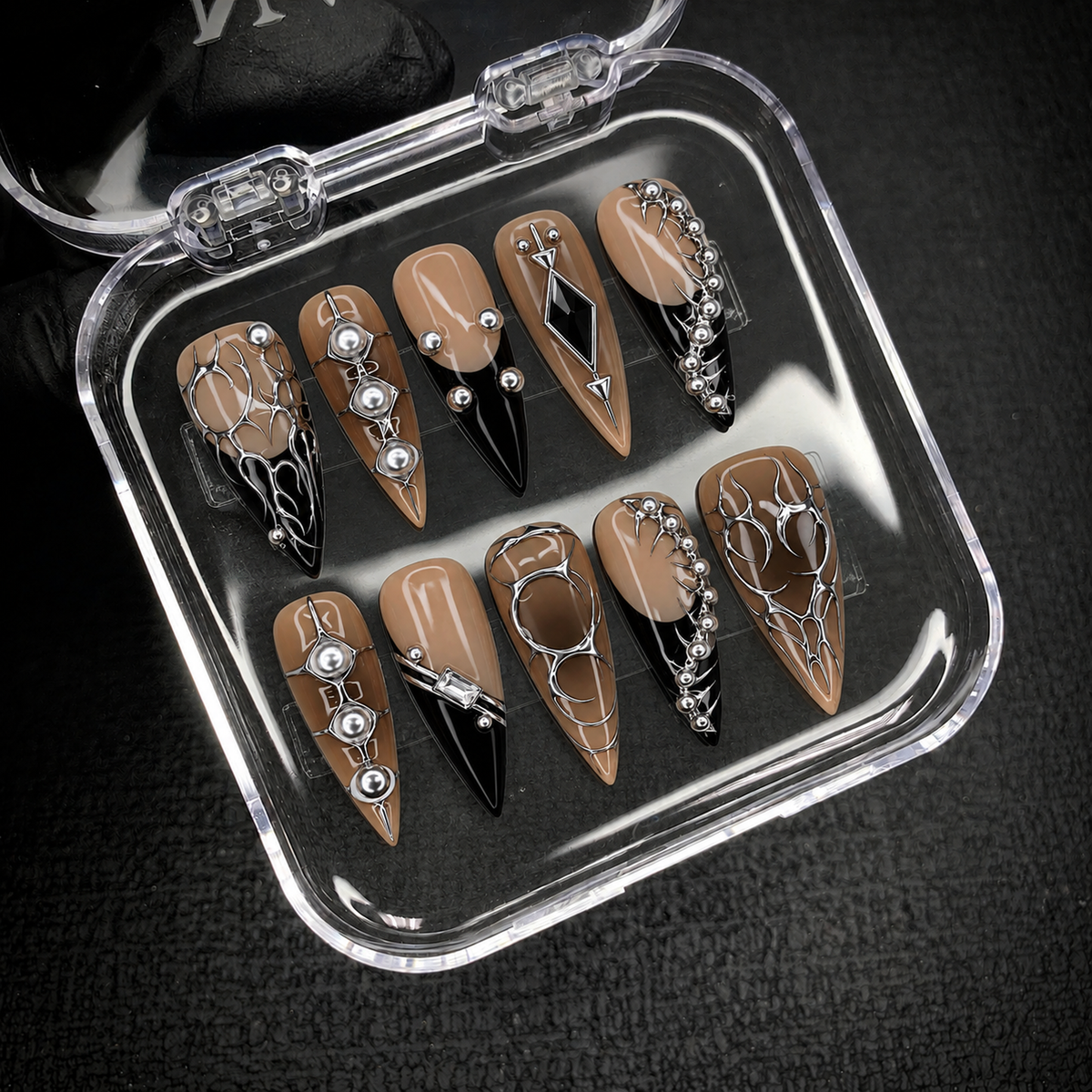 Open clear case on dark textured fabric displaying ten coordinated taupe and black nails with silver-tone ornament and geometric accents.