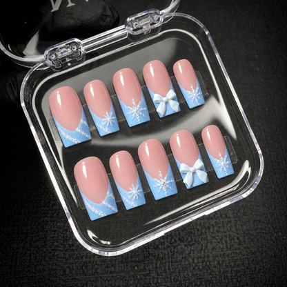 Open clear case showing ten pink and blue press-on nails with snowflake designs and white bow accents.