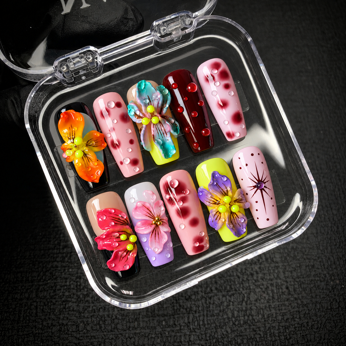 Open clear case on a dark textured background holding ten coordinated nails with sculptural flowers, burgundy spots, and glossy droplet details.