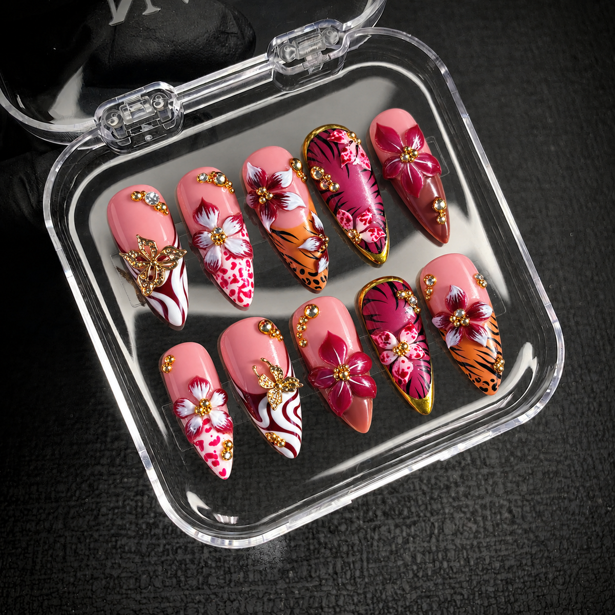Open clear case holding ten pink floral and animal-print nails with butterfly ornaments and gem accents on a dark textured surface.