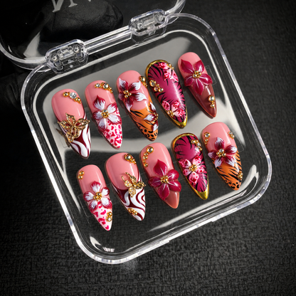 Open clear case holding ten pink floral and animal-print nails with butterfly ornaments and gem accents on a dark textured surface.