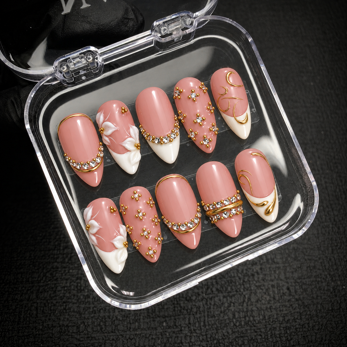 Open clear nail case holding ten coordinated blush-pink, white, and gold-tone decorated nails on a dark textured surface.