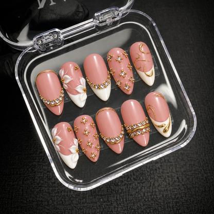 Open clear nail case holding ten coordinated blush-pink, white, and gold-tone decorated nails on a dark textured surface.